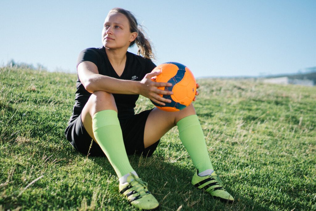 Na zdjęciu widzimy sportową kobietę, która siedzi na trawniku trzymając w rękach piłkę do footballu. Kobieta ma na sobie wygodny strój sportowy i sportowe korki na nogach, które zapewniają jej stabilność i przyczepność podczas wykonywania akrobacji z piłką. Jej uśmiechnięta twarz i pewna poza świadczą o tym, że czerpie radość i satysfakcję z uprawiania sportu. Tło zdjęcia stanowi zielony trawnik, który idealnie wpisuje się w klimat sportowej aktywności na świeżym powietrzu. To inspirujące zdjęcie pokazuje, że sport to nie tylko zdrowie i dobre samopoczucie, ale także radość i przyjemność z uprawiania ulubionej aktywności fizycznej